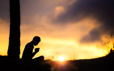 Silhouette of young male christian sitting and praying to god with light of sunset background,...