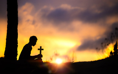 Obraz na płótnie Canvas Silhouette of young male christian sitting and holding a cross for blessing from god with light of sunset background, christian hope concept.