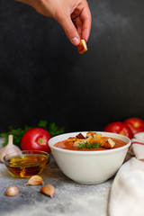 Traditional summer cold tomato soup gazpacho. Tomatoes, garlic, basil, olive oil. Woman hand holding crouton. Mediterranean, Spanish cuisine. Front view. Black background. Copy space