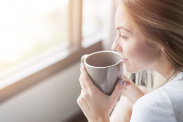 Relaxation time. Close up of cheerful blonde girl drinking coffee and looking through the window