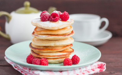 stack of baked homemade pancakes with raspberries, sprinkled with powdered sugar and tea utensils in the background, selective focus