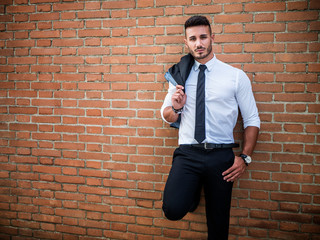 Portrait of stylish young man wearing business suit, standing in modern city setting, leaning against brick wall