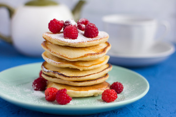 stack of baked homemade pancakes with raspberries, sprinkled with powdered sugar and tea utensils in the background, selective focus