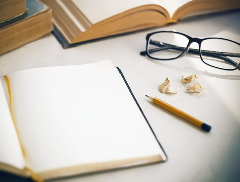 On the white table there are old books, an open diary with a yellow bookmark for notes, shavings from a sharpened, a yellow pencil and glasses in a black frame. Everything is ready for study and work. - Powered by Adobe