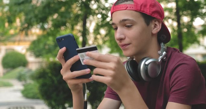Teen makes online purchases using a credit card and mobile phone.  Smiling young man makes online shopping using cell phone with bank card, outdoors. Boy uses smartphone and credit card. Real time.