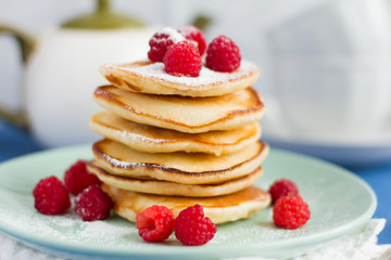 stack of baked homemade pancakes with raspberries, sprinkled with powdered sugar and tea utensils in the background, selective focus