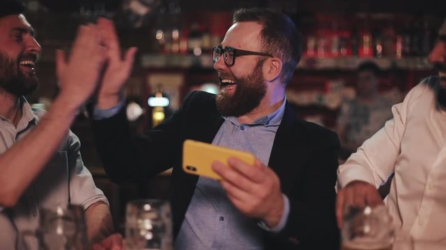 Three old friends watching video on smartphone and drinking draft beer in the sport bar. Friends laughing and discuss the news in beer pub.