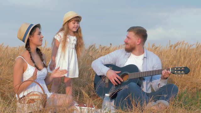 countryside family leisure, young man plays guitar while his beautiful wife and cute little daughter dance laughing at picnic with milk and buns in grain harvest field of golden wheat shining by