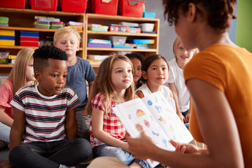 Group Of Elementary School Pupils Sitting On Floor Listening To Female Teacher Read Story - obrazy, fototapety, plakaty