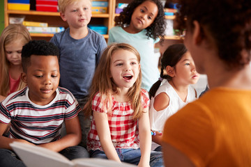Group Of Elementary School Pupils Sitting On Floor Listening To Female Teacher Read Story