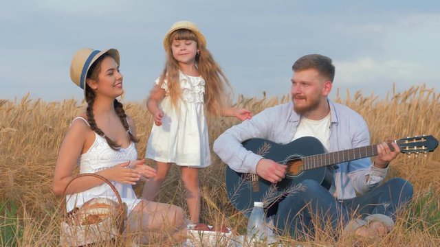 family fun in nature, young cheerful daddy plays guitar when his beautiful wife and little pretty daugther clap hands happily dancing during picnic in shining by sunbeams wheat harvest field