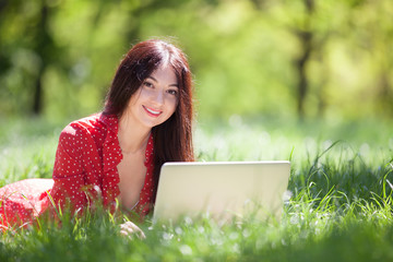 Young woman with white laptop in the park. Beauty nature scene with colorful background, trees at summer season. Outdoor lifestyle. Happy smiling woman with computer lay on green grass