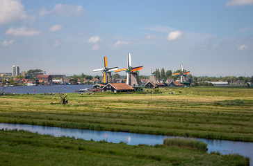 Fototapeta premium Zaanse Schans, Netherlands - 06/14/2019: Old dutch windmills and river in historical village. Holland mills in field panoramic view. Rural holland landscape. Wind energy concept. 