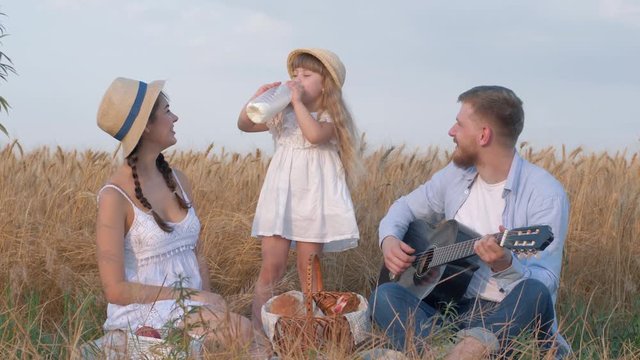 happy family moments, little cute kid girl drinks tasty milk from bottle while her beautiful parents laugh and young dad play guitar during outdoor picnic in grain oat field at harvest autumn time