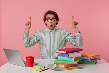 Young surprised student in glasses, sits by the table and working with laptop, looks at the camera and wants draw you attention to copispee over his head, isolated over pink background.