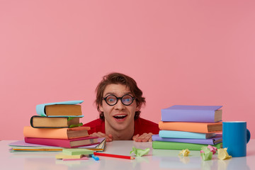 Portrait of positive man in glasses wears in red t-shirt,hiding at the table with books, looks at the camera and smiling, looks cheerful, isolated over pink background.