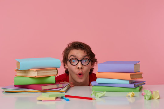 Portrait of happy amazed man in glasses wears in red t-shirt, hiding at the table with books, looks at the camera with surprised expression, looks cheerful, isolated over pink background.