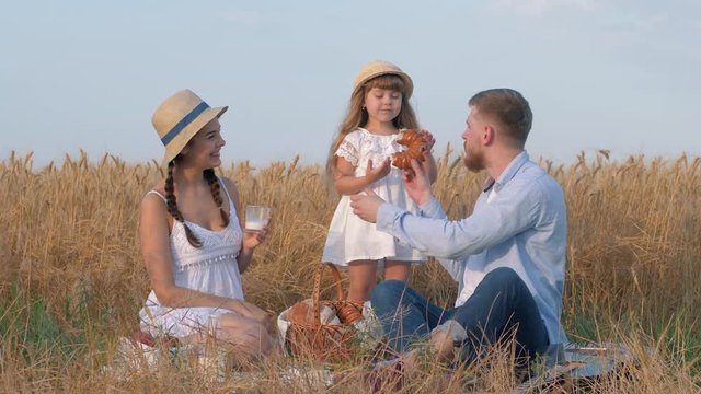 happy family portrait, young father bites sweet bun from his daughter hands and smiles to pretty woman sitting in open air sunny gold grain meadow picnic during harvest autumn season
