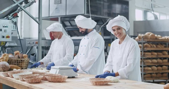 Attractive woman baker and her colleagues bakers working fast in a bakery industry forming pieces of dough process of making bread in a bakery commercial kitchen