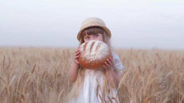 happy farmer daughter, little cute child girl eats tasty freshly baked rooty and smiles happily staying in golden barley field of good harvest bread season against sky