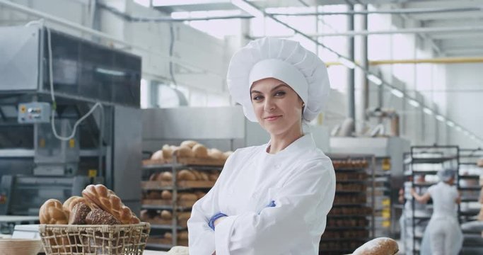 In front of the camera charismatic baker woman in a industrial kitchen smiling large and looking stray to the camera background big industrial bakery machine