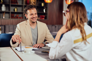 Two multicultural smiling colleagues dressed smart casual sitting in restaurant and discussing about project.