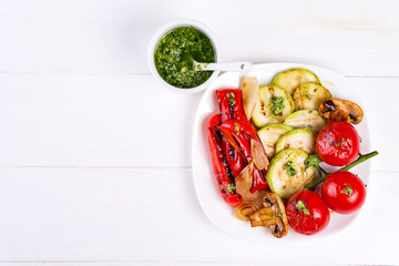 Grilled vegetables on a plate with pesto on white wooden background, flat lay