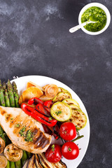 Chicken breast grill with bbq vegetables and pesto sauce in a plate on a concrete background, flat lay,