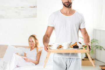 panoramic shot of man holding food tray with breakfast in bedroom