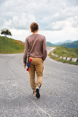 Fototapeta premium Young stylish man with long hair gathered in a ponytail and in sunglasses walking with a longboard in his hands on a country asphalt road. Mountains in the evening