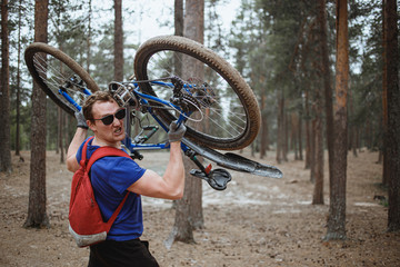 Handsome strong man looking at camera while walking in forest and holding bike over head. Energy...