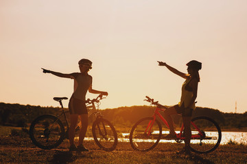 Two silhouettes of women with bike standing on lakeside and talking each other at the sunset