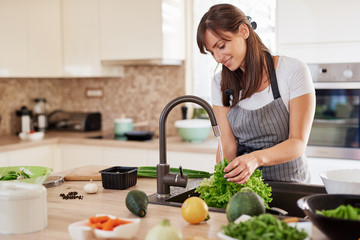 Cheerful attractive Caucasian woman in apron washing salad in kitchen. Dinner at home concept.