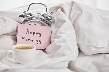 Fototapeta na wymiar coffee in white cup near silver alarm clock with happy morning lettering on sticky note in bed