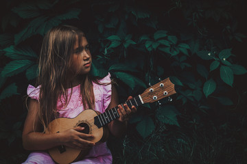 Obraz na płótnie Canvas Beautiful young girl with blond hair wearing pink dress playing ukulele and dreaming on dark nature background. Grape leaves. Cutie lady learns play guitar concept