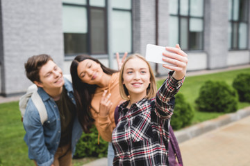 happy teenagers holding smartphone, taking selfie and smiling outside