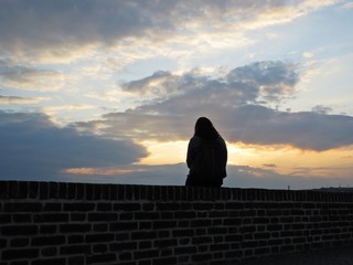 One Single Woman Silhouette Sitting on Brick Wall during Sunset with Cloudy Sky in Background