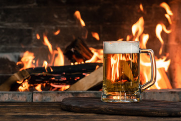 Beer in a mug on a wooden table on a burning fire in a fireplace background.
