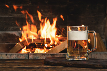 Beer in a mug on a wooden table on a burning fire in a fireplace background.