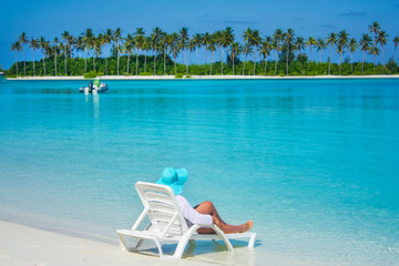 women in a blue hat lies on a white lounger on the beach in the Maldives, looks at a beautiful paradise landscape with blue sky, water and an island with palm trees