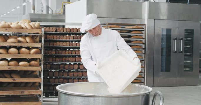 Bakery factory old man chef preparing the dough in a industrial container he add some flour while in background bakery workers transported the baked bread. 4k