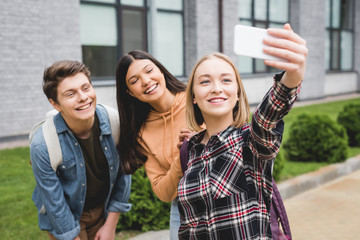 cheerful teenagers holding smartphone, taking selfie and smiling outside