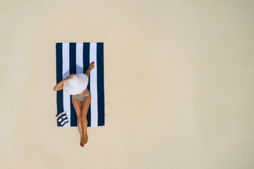 Summer holiday fashion concept - tanning girl wearing sun hat at the beach on a white sand shot from above.Top view from drone. Aerial view of slim woman sunbathing lying on a beach in Maldives.