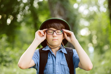 Portrait of little explorer in forest. Boy traveler in helmet play in the park. Happy child in glasses go hiking with backpack in summer nature. Dream concept.