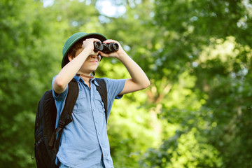 Portrait of little explorer with binoculars in forest. Boy traveler in helmet play in the park. Happy child go hiking with backpack in summer nature. Dream concept.