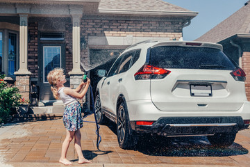 Cute preschool little Caucasian girl washing car on driveway in front house on sunny summer day. Kids home errands duty chores responsibility concept. Child playing with hose spraying water.
