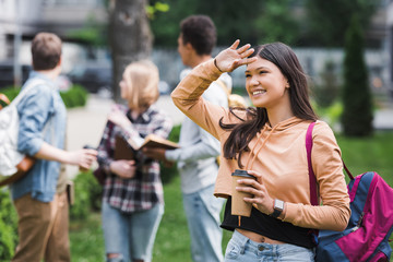 smiling and brunette teenager holding paper cup with coffee and looking away