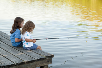 children are fishing from wooden pier on a lake. two cute sisters have fun fishing. Family leisure activity during summer sunny day. Active family time on nature. Hiking and adventure with little kid 