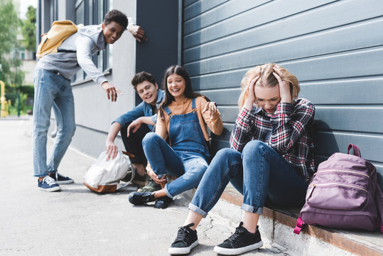 smiling friends bulling and pointing with fingers at sad blonde teen