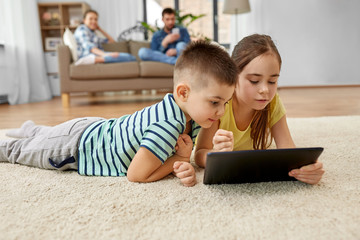 childhood, technology and family concept - brother and sister with tablet computer lying on floor...